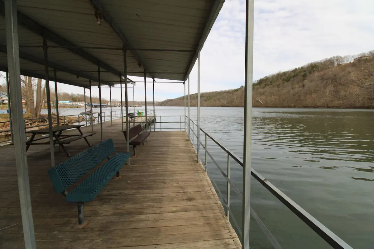 A wide waterfront view featuring a dock structure, comfortable benches for sitting, and beautiful water vistas. The scene is ideal for fishing, picnicking, or simply watching the sunrise over the lake. The mood is peaceful, inviting relaxation and storytelling by the water’s edge.
