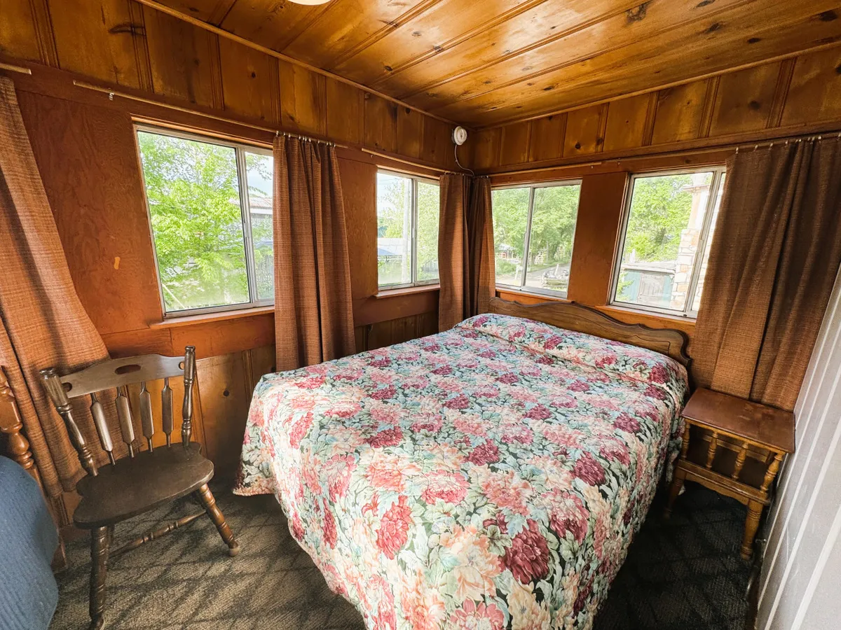 A sunlit bedroom with a cozy bed draped in a floral quilt, a wooden chair, and large windows framing views of the outdoors. The simplicity of the decor and the warmth of the natural light create a peaceful, intimate atmosphere ideal for relaxation.