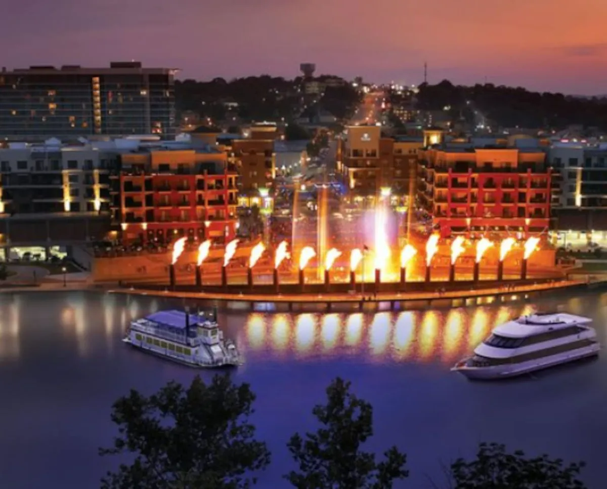 This vibrant exterior scene shows colorful buildings lining the waterfront, lively fountains with flames, and boats gently floating on the water. The festive mood is underscored by people dining near the water and the potential for evening events like fireworks shows.