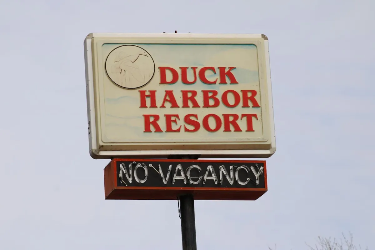 An exterior shot featuring the Duck Harbor Resort sign with a 'No Vacancy' indication, surrounded by greenery and hints of the lakeside setting. The nostalgic feel conjures memories of classic family vacations and lakeside getaways.