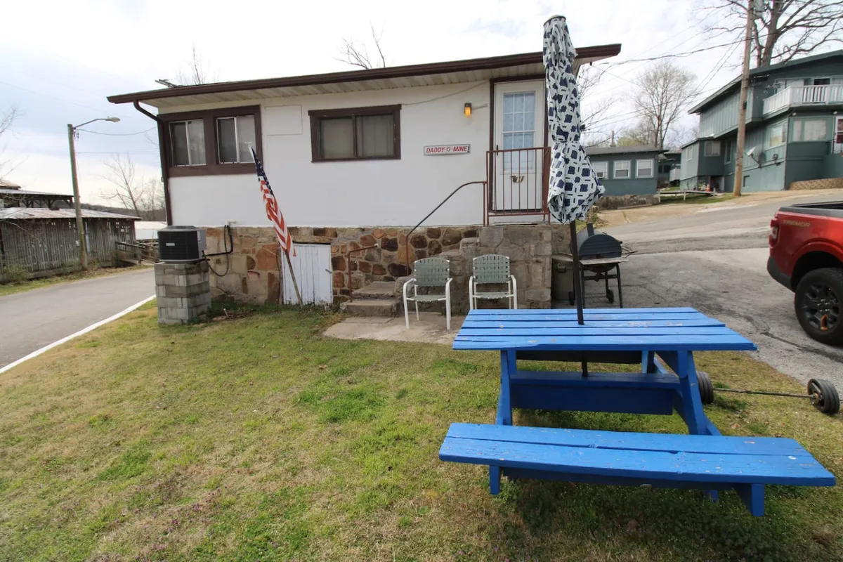 A blue picnic table with an umbrella overlooks the lake, surrounded by a stone wall, flag, and comfortable chairs. A barbecue grill is nearby, creating a welcoming space for outdoor dining and social gatherings.