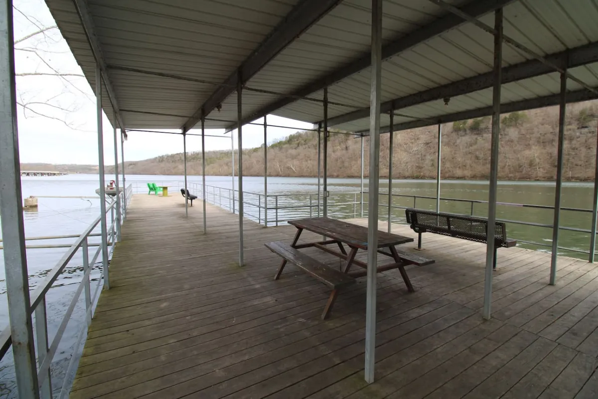 A waterfront scene showing a wooden dock, picnic table, green chairs, and a peaceful river view. The dock is ready for fishing or boating, and the setup invites easy access to water-based activities.