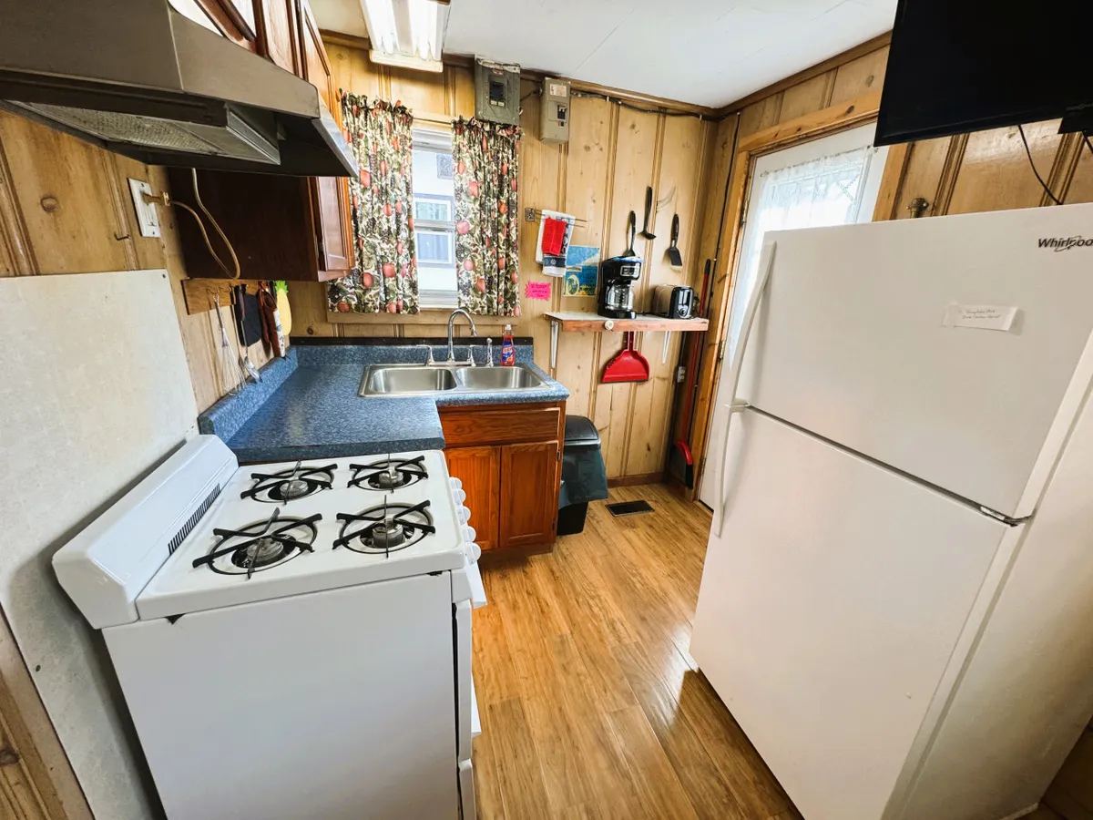 This interior image showcases a kitchenette with a stove, sink, refrigerator, and open shelving, all accented by homey curtains. Essential cookware is visible on the shelves, and the inviting setup is clearly designed for meal preparation and casual dining.