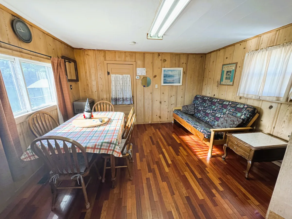 A welcoming living and dining area featuring a wooden dining table with a checkered tablecloth, a cozy sofa with patterned upholstery, wooden flooring, and a large window letting in natural light. The inviting space encourages both relaxation and social gatherings, with a clear view to the outdoors.