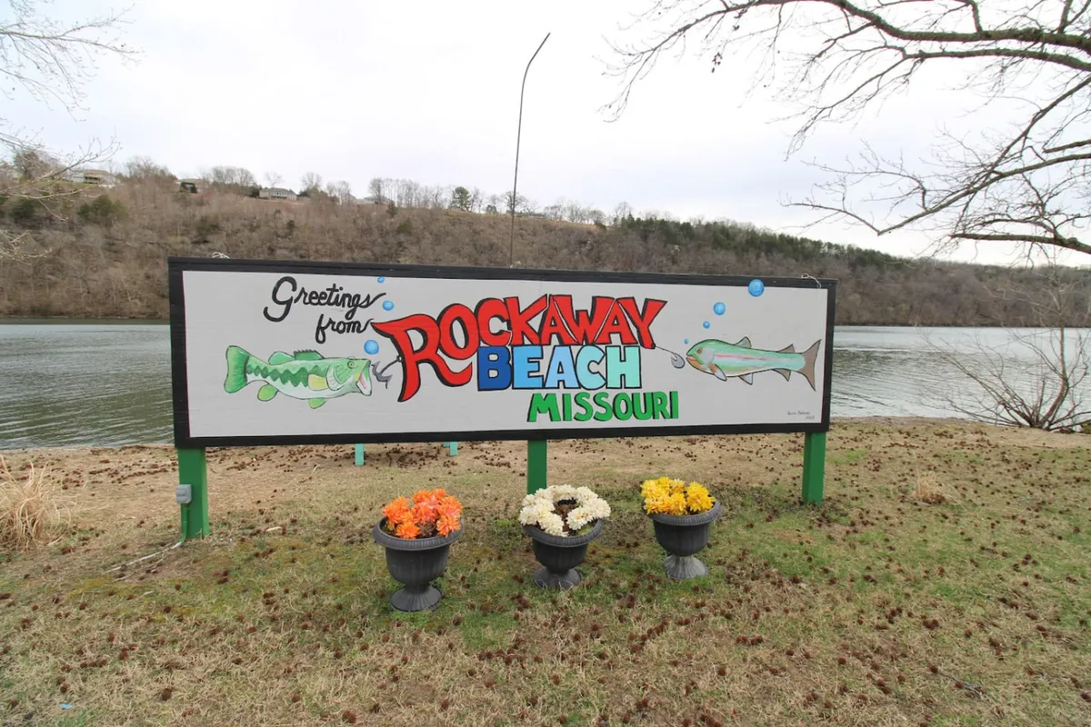 The image shows the exterior of a cozy lakeside cabin at Rockaway Beach, Missouri. The welcoming sign, colorful flower pots, and a beautiful view of Lake Taneycomo in the background set a friendly, inviting mood. The scene hints at outdoor activities like fishing, picnicking, and nature walks, all within steps of the cabin.