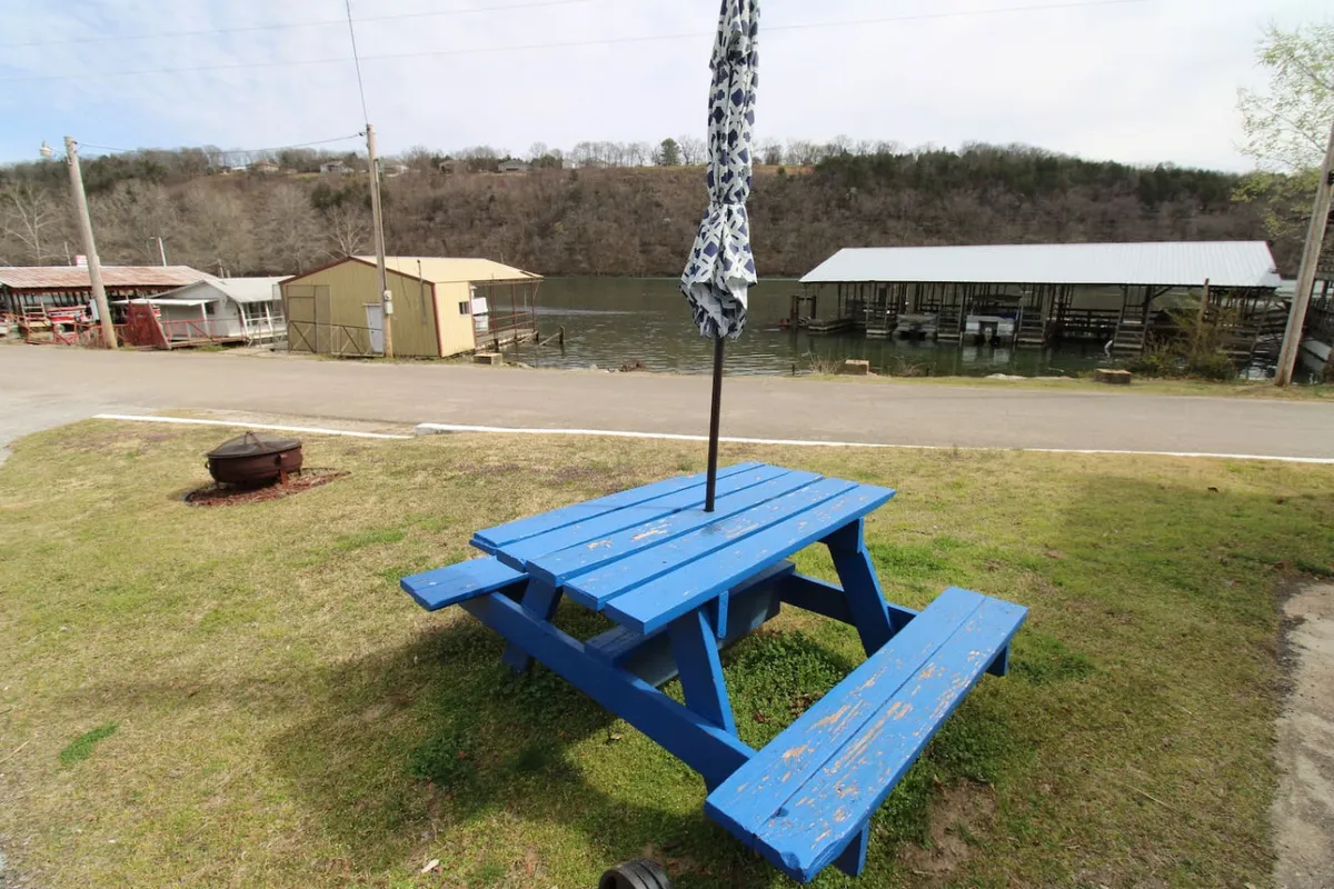 A vibrant blue picnic table and umbrella sit right by the water's edge, offering a clear lakeside view. The table is set up for dining, relaxation, or enjoying the sunset over the lake.