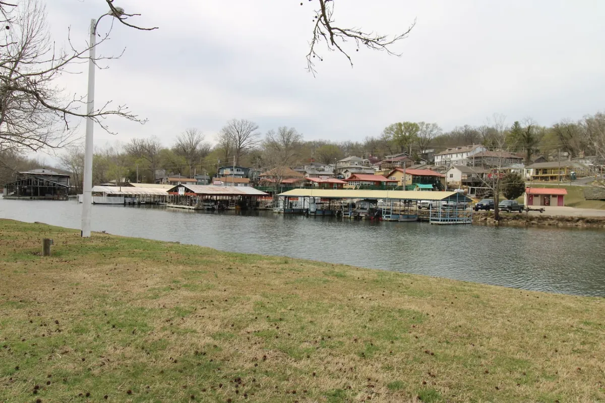 A dock with boats stretches over the water, with waterfront houses and lush greenery lining the shore. The sun sets in the background, casting a warm glow. The image evokes leisure activities like evening boat rides, waterfront picnics, and barbecues by the shore, tying into the retreat's lakeside lifestyle.
