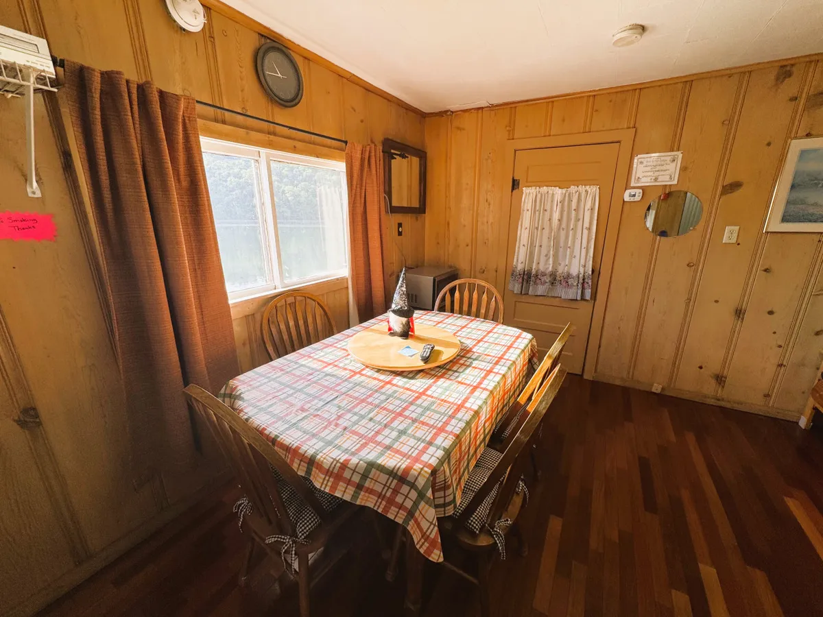A dining space with a checkered tablecloth on a dining table, surrounded by wooden chairs, and a window with curtains providing a view outward. A wall clock and warm decor further emphasize the room’s welcoming feel.