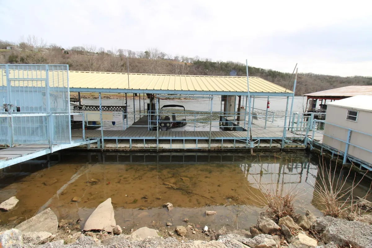 A serene scene featuring a boathouse and dock extending over the tranquil waters of Lake Taneycomo. The surrounding landscape is lush and inviting, with ample space for fishing or relaxing by the water’s edge. The dock area is perfect for catching fish or launching a boat, while an adjacent area is set up for picnics and outdoor grilling.