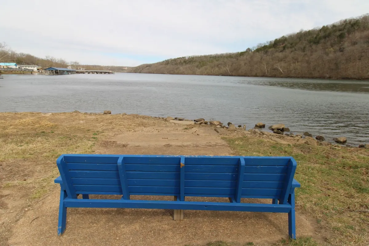 A tranquil lakeside scene featuring a blue wooden bench positioned by the calm water, surrounded by lush trees. The bench invites quiet reflection, while the peaceful lake hints at opportunities for relaxing, fishing, or simply soaking in nature. The overall ambiance exudes solitude and serenity—perfect for those seeking a peaceful retreat.