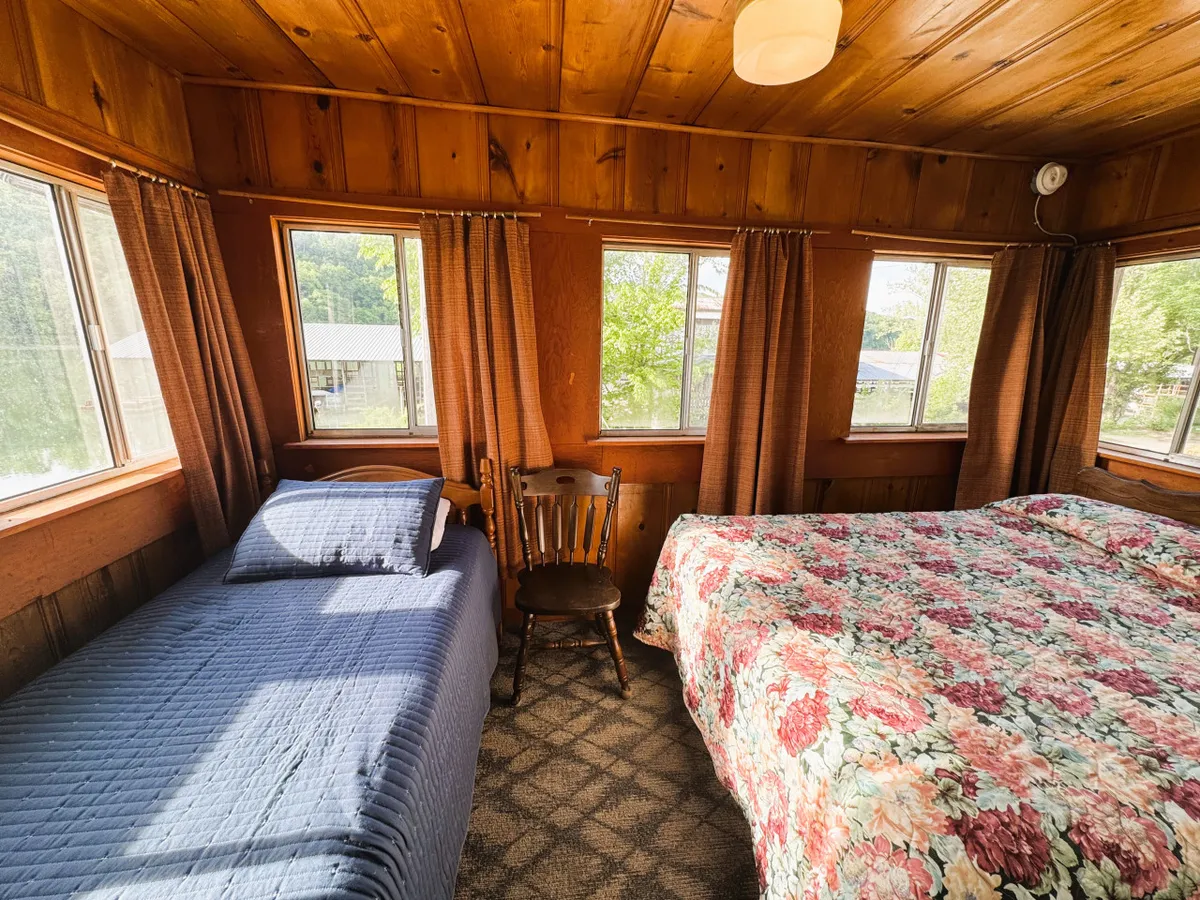A bright, welcoming bedroom with two comfortable beds, a wooden chair, and large windows that let in abundant natural light. The room is styled simply but warmly, inviting relaxation and offering views of the greenery outside. The layout is practical and cozy, ideal for restful nights after a day of adventure.