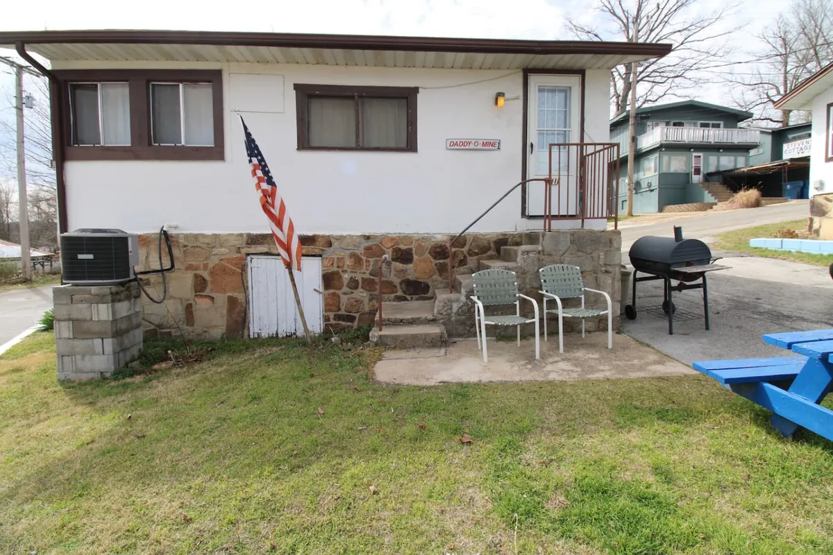 A close-up of a barbecue grill with two chairs beside a stone wall and an American flag fluttering in the background, all under a sunny sky. The setup invites guests to relax and socialize while enjoying grilled meals.